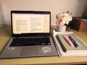 From familiar to unfamiliar. Ashley only just read Darkness, Set Us Free, after having a two month break from the manuscript. In the picture is her laptop, notebook and writing utensils, with roses in a vase for decoration.